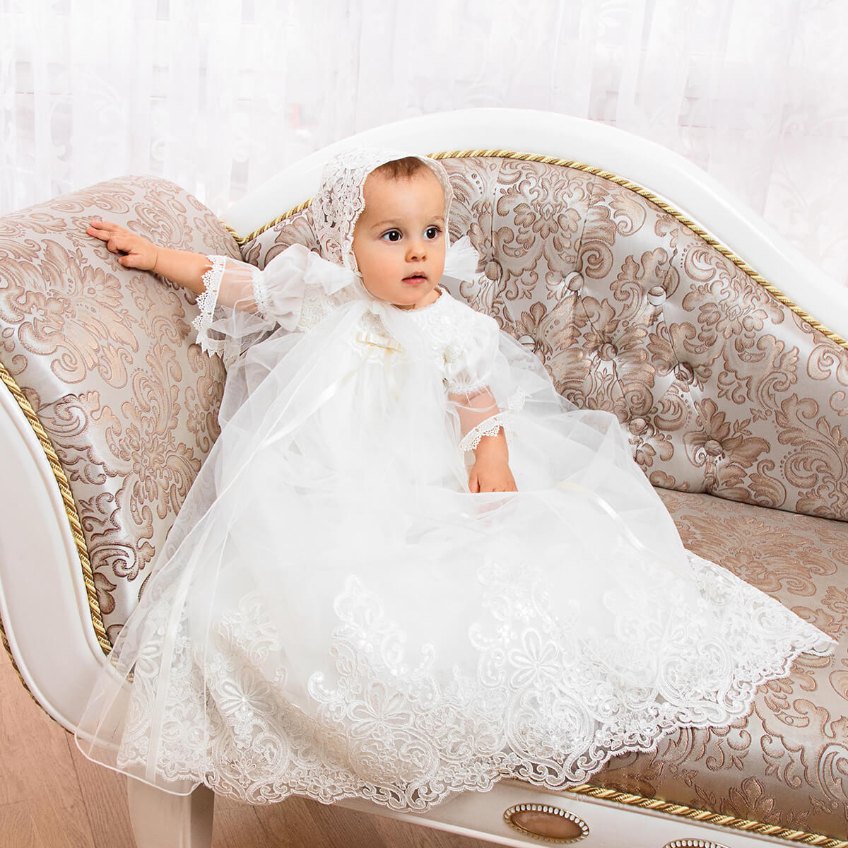 Baby in a Christening Gown with Train for Girls  sitting on an ornate couch.
