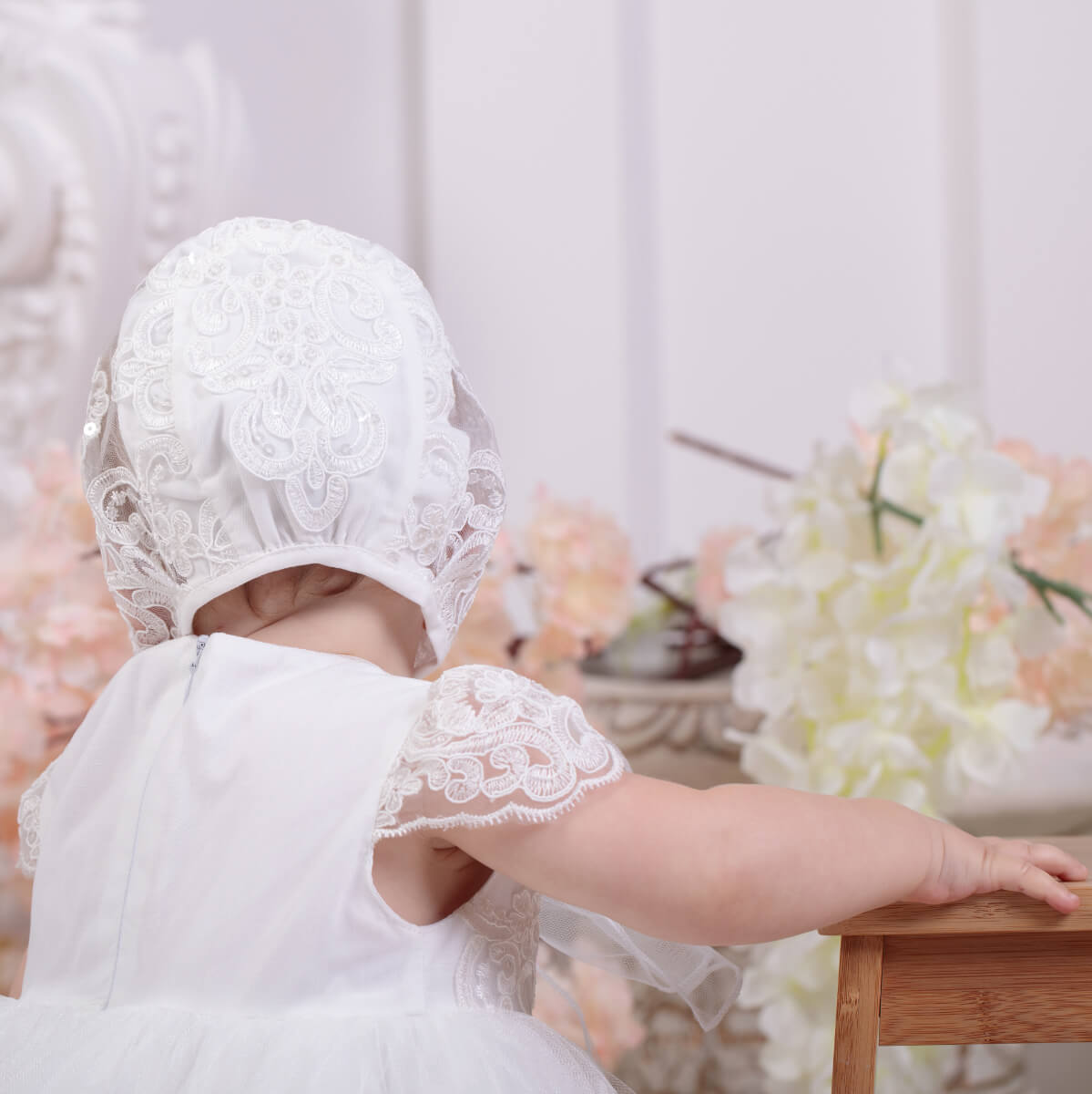 Baby in a Lace Baptism Bonnet and dress sitting at a table with a floral background