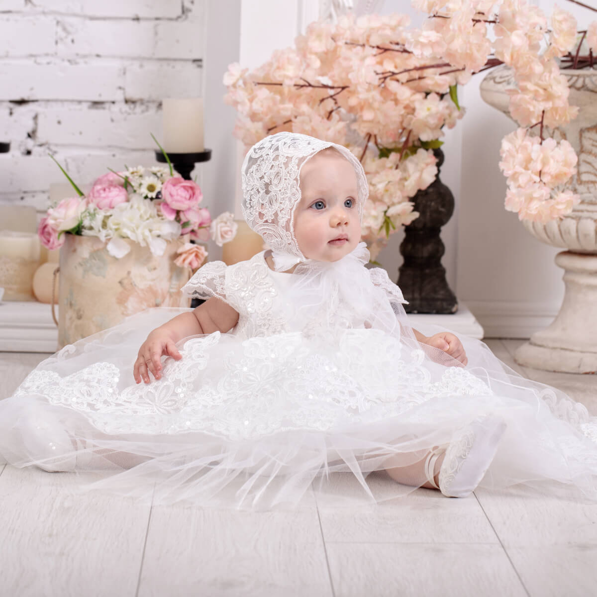 Baby in a Lace Baptism Dress with Train and bonnet sitting on a light-colored surface with floral decorations in the background.