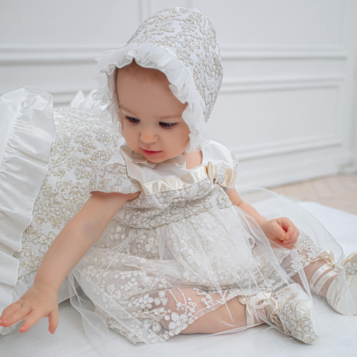Baby in a  Baby Baptism Dress and bonnet sitting on a white surface.