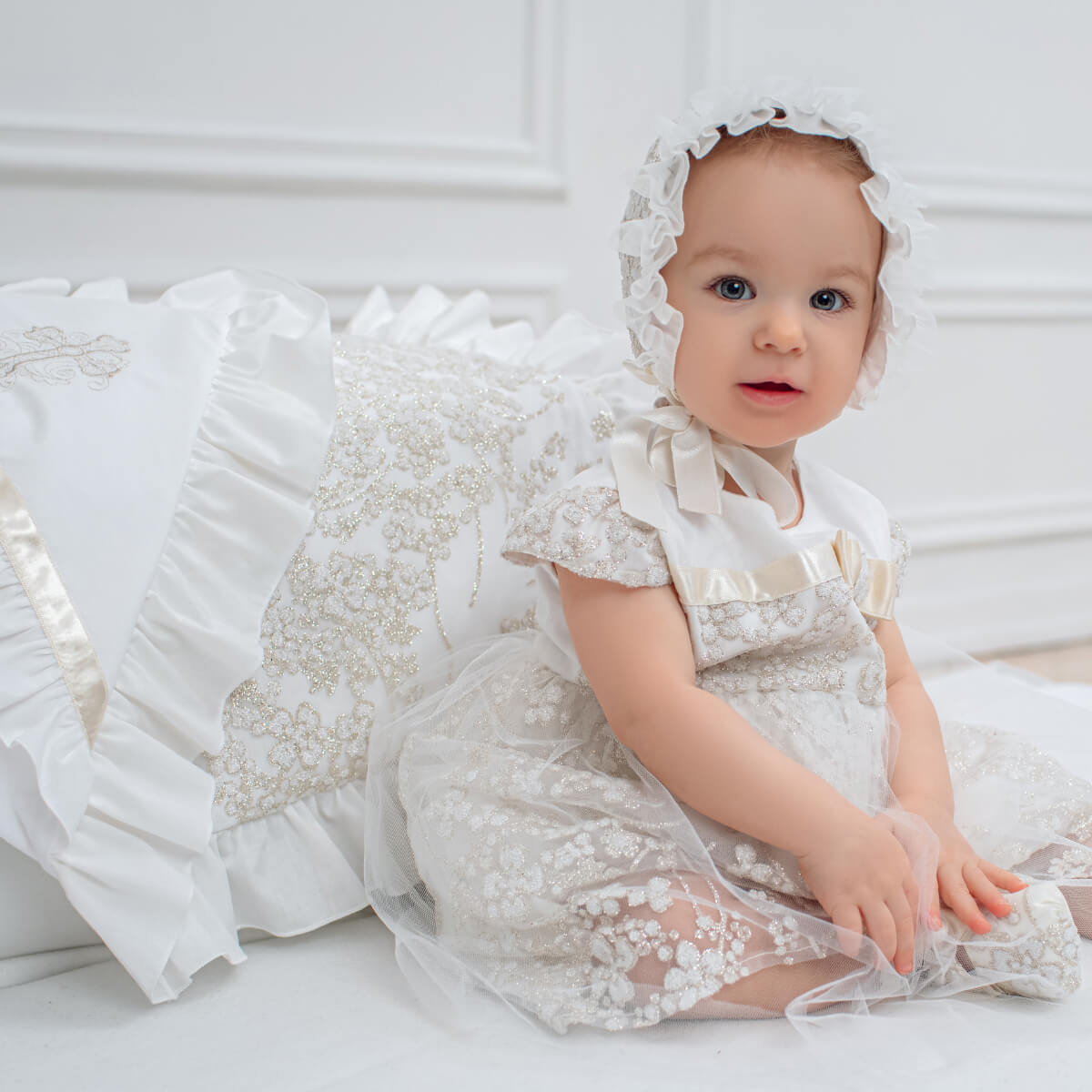 Baby in a  Baby Baptism Dress and bonnet sitting on a white surface.