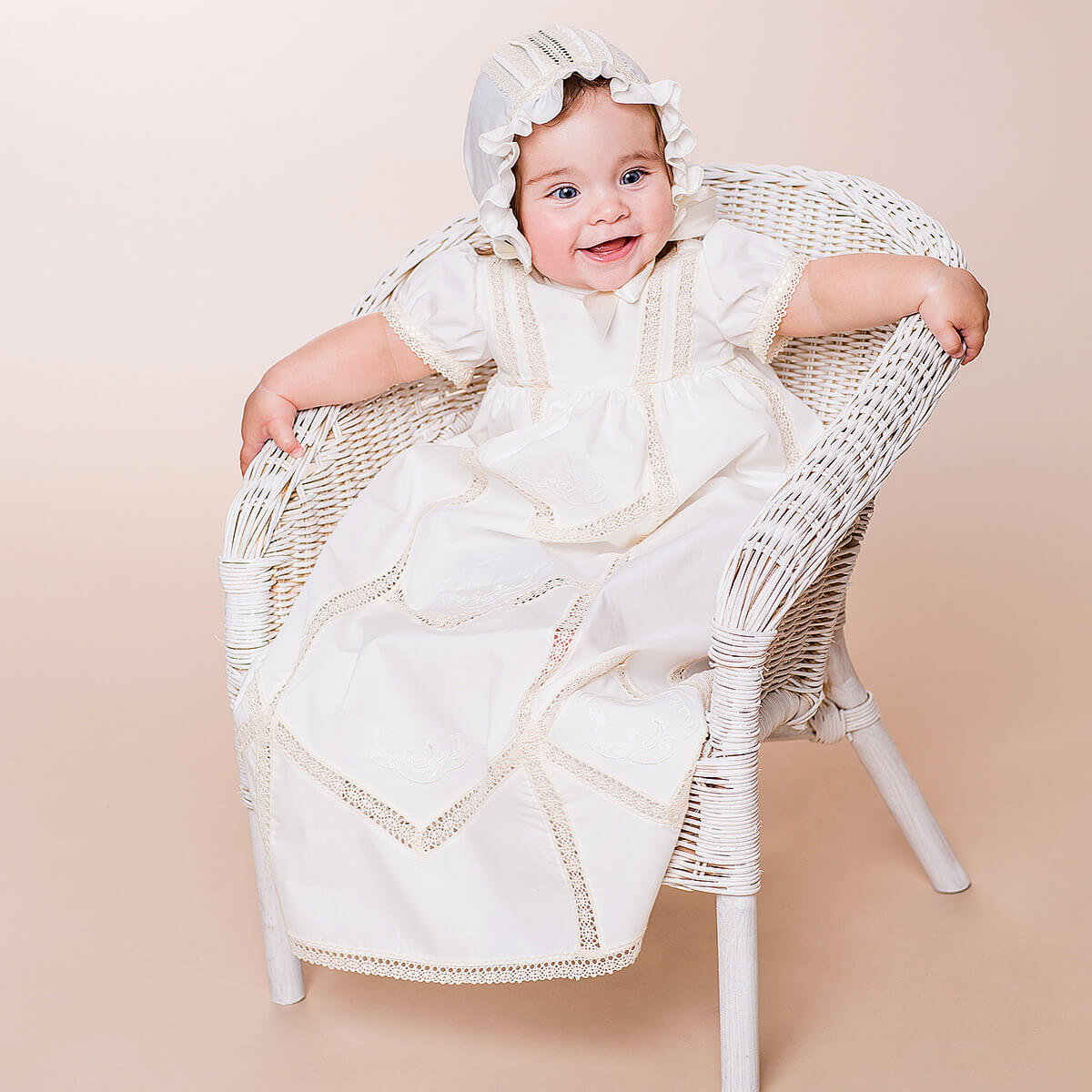 Baby in a white dress and Lace Baptism Bonnet sitting on a wicker chair against a beige background