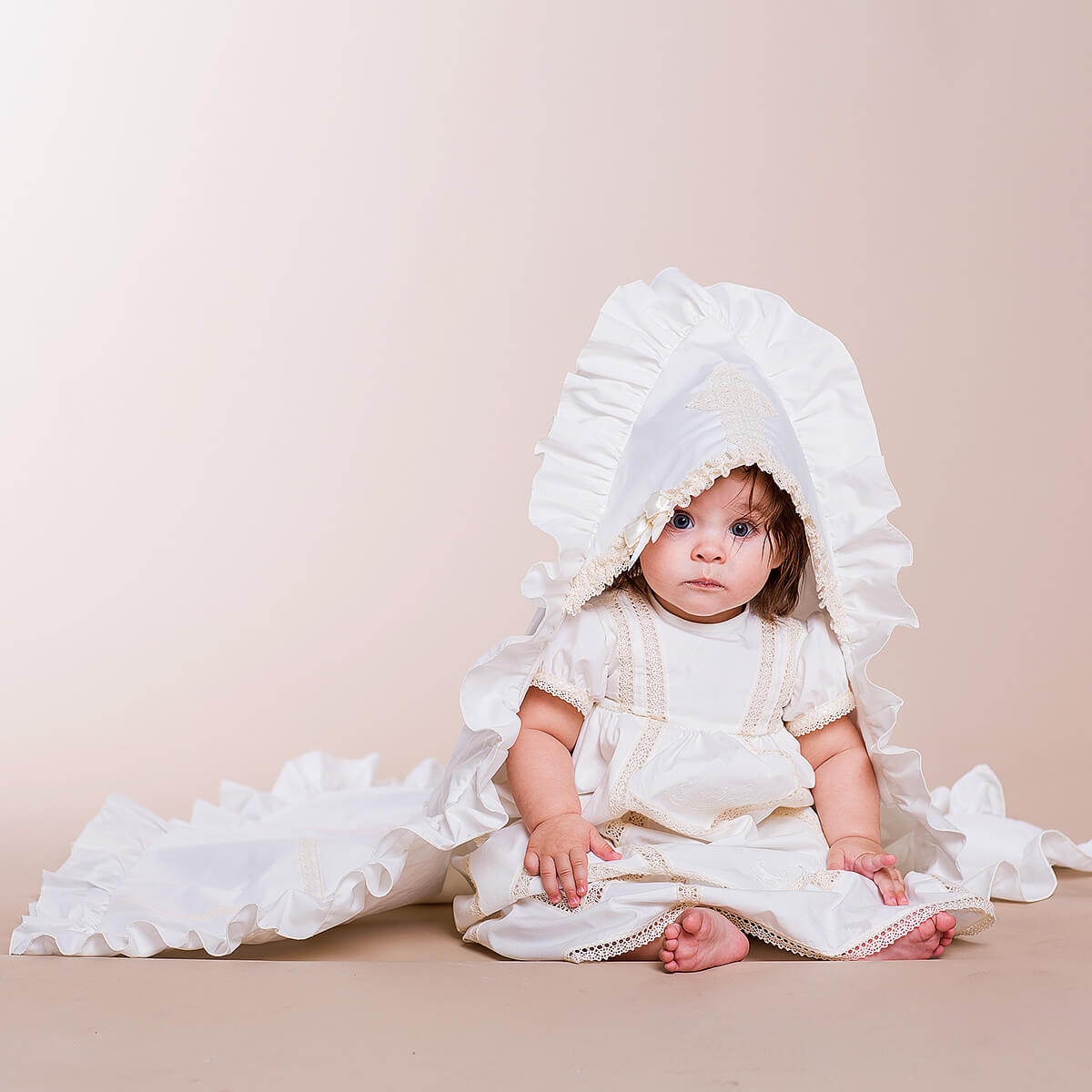 Baby in a white dress with a large ruffled baptism blanket on a beige background