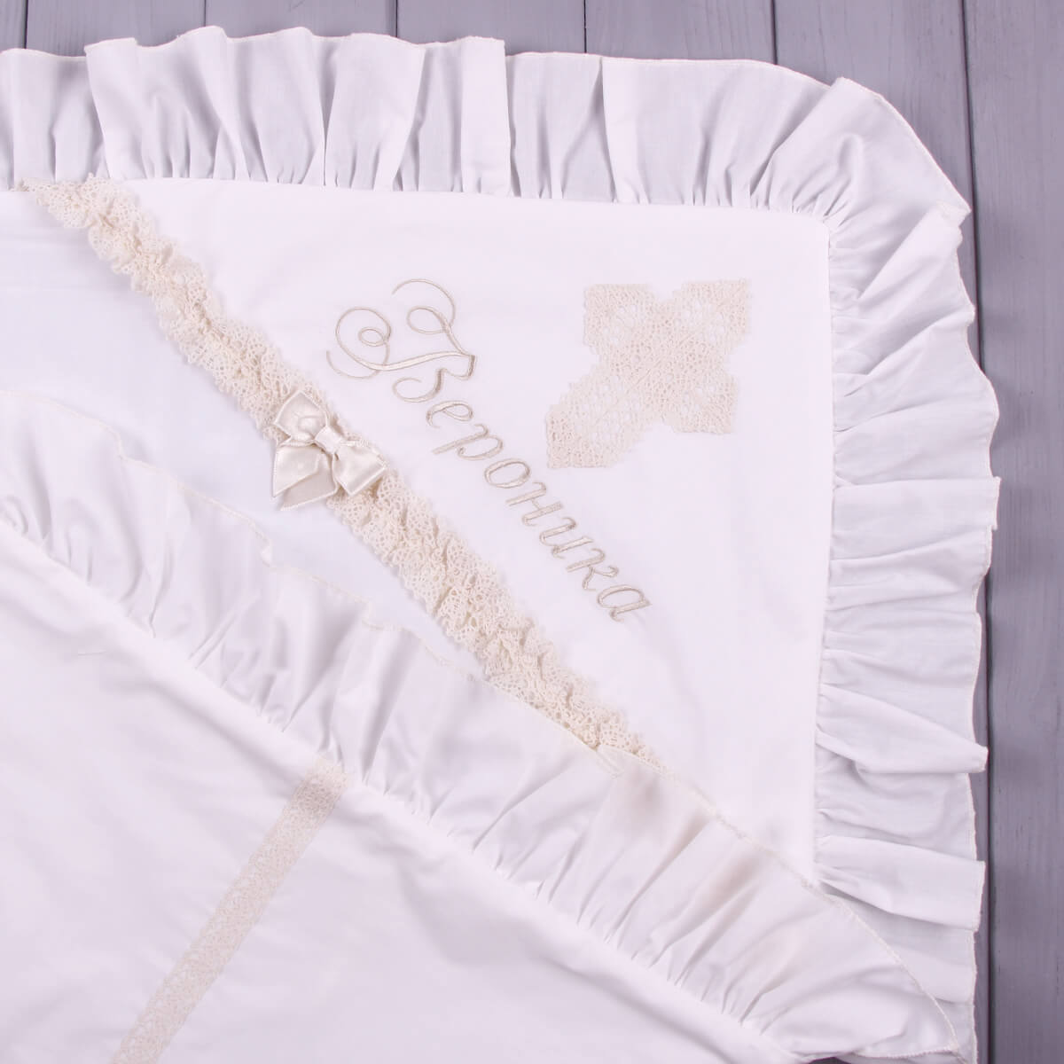 Ivory baptism blanket with lace trim and embroidered name on a wooden surface
