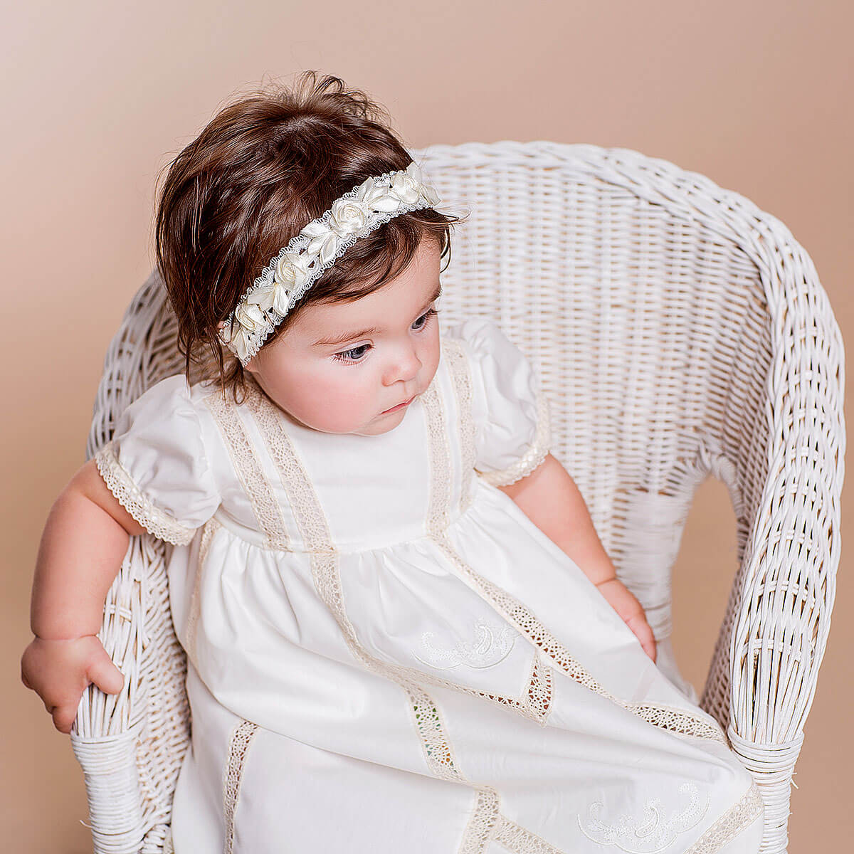 Baby in a  Christening Gown for Girls with lace details sitting on a wicker chair against a beige background