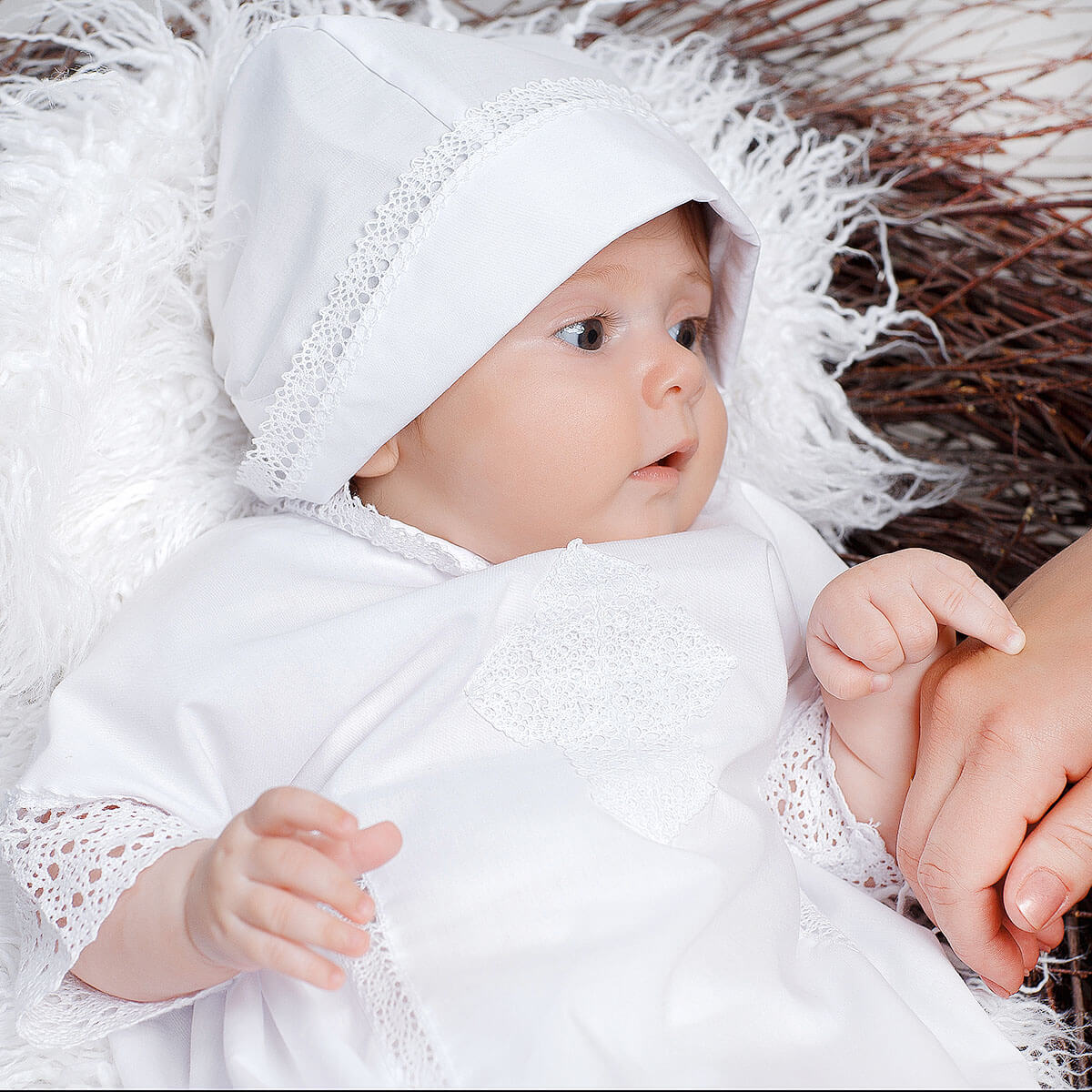 Baby in a white outfit with a Baptism Bonnet sitting on a textured surface.