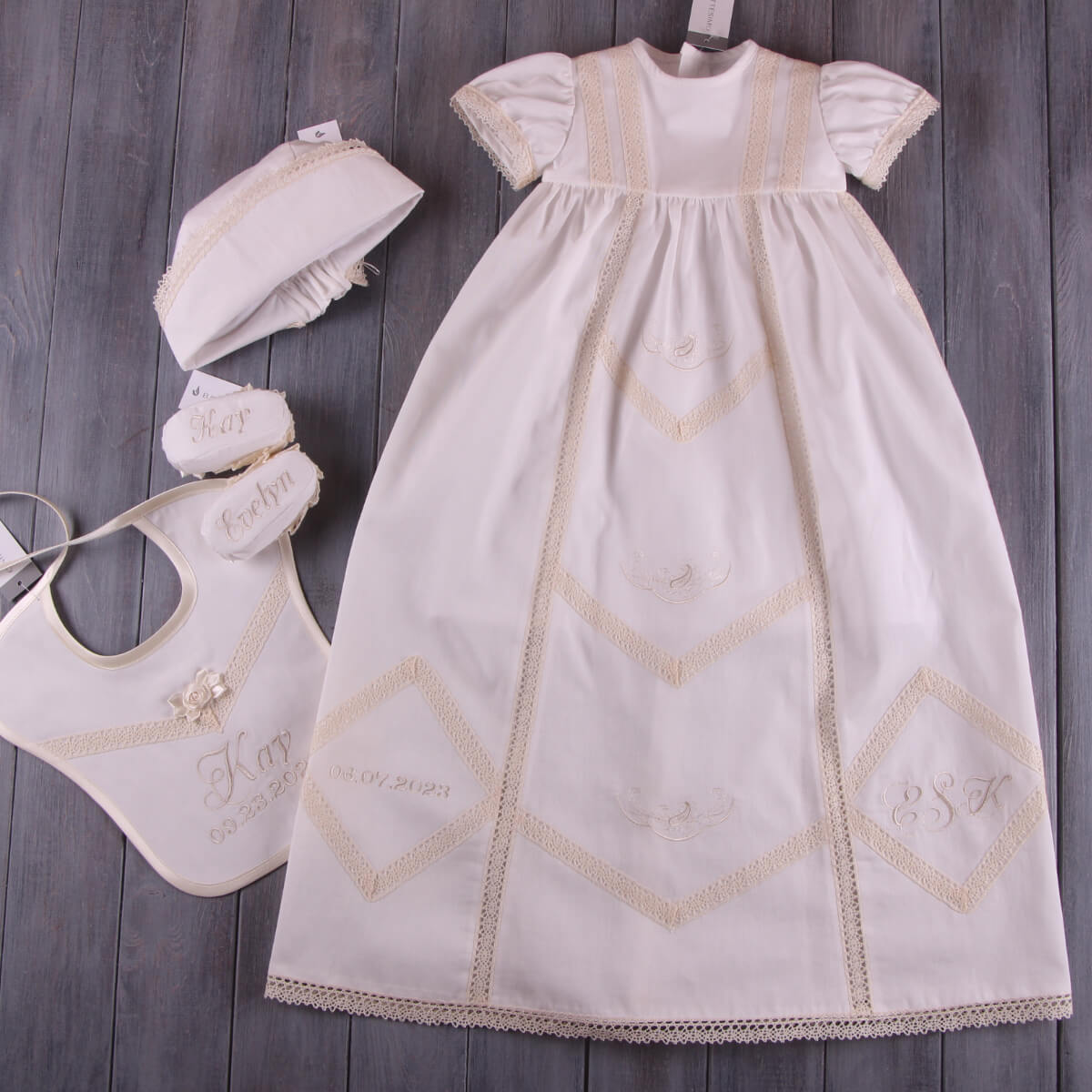 White baby dress with gold embroidery, hat, and  Newborn Bib on a wooden surface