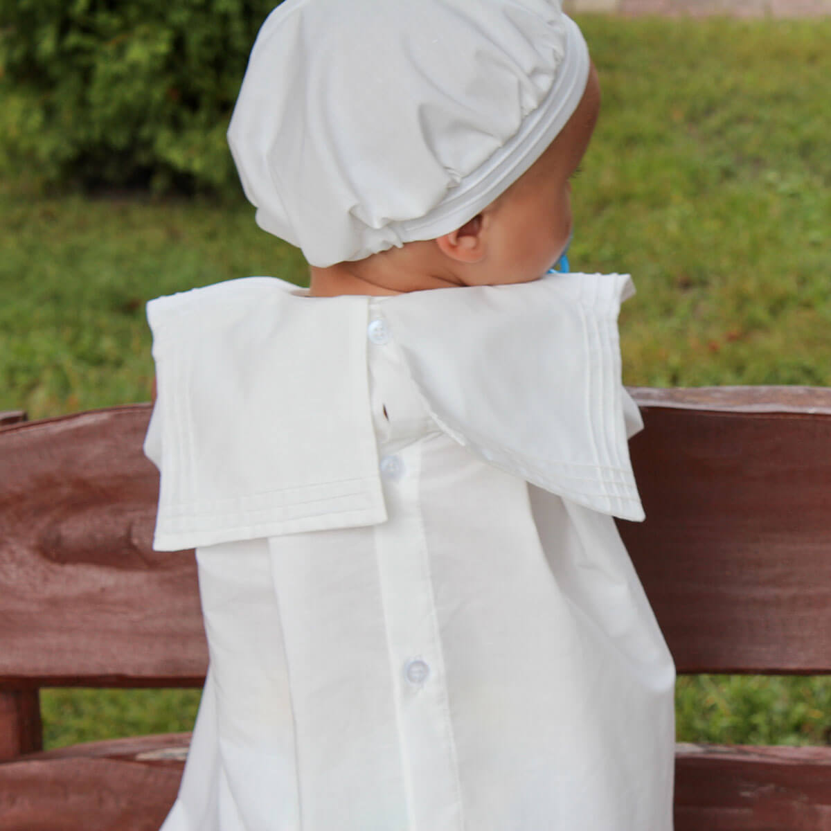 Baby in a christening outfit sitting on a wooden bench outdoors.