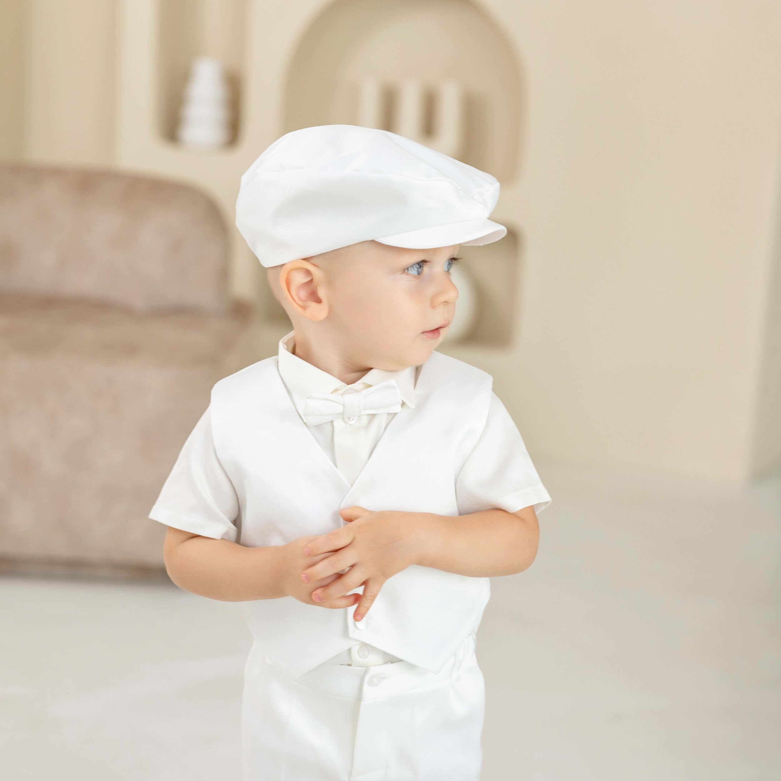 Baby in a Baby Boy Christening Suit  with a bow tie and cap, standing indoors.