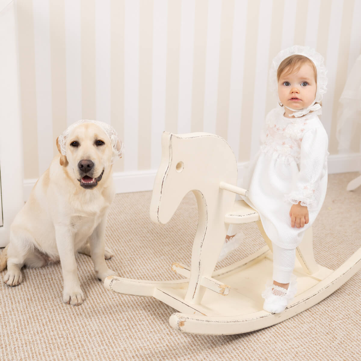 Baby in a Newborn Coming Home Dress sitting on a rocking horse next to a dog.