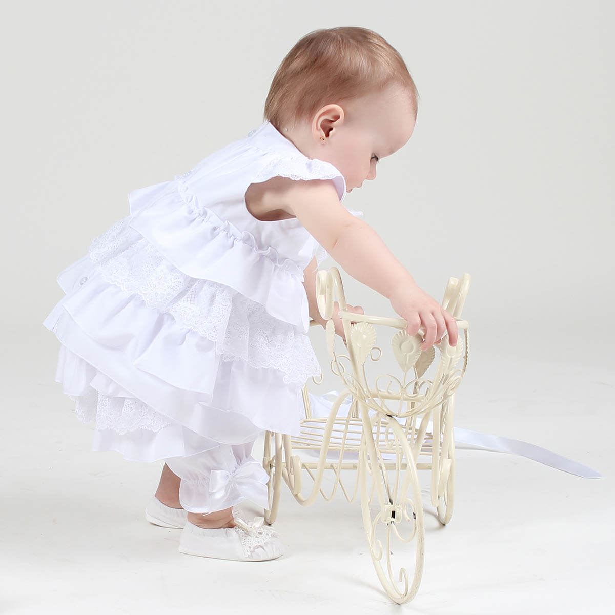 Christening Lace Dress playing with a small white chair on a plain background